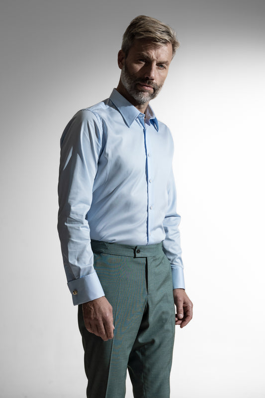 Moden kaukasisk mand i en lyseblå skjorte og grønne bukser, stående mod en hvid baggrund