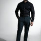 Stilfuld mand med skæg i en sort skjorte og mørke bukser, udstråler selvtillid mod en enkel baggrund