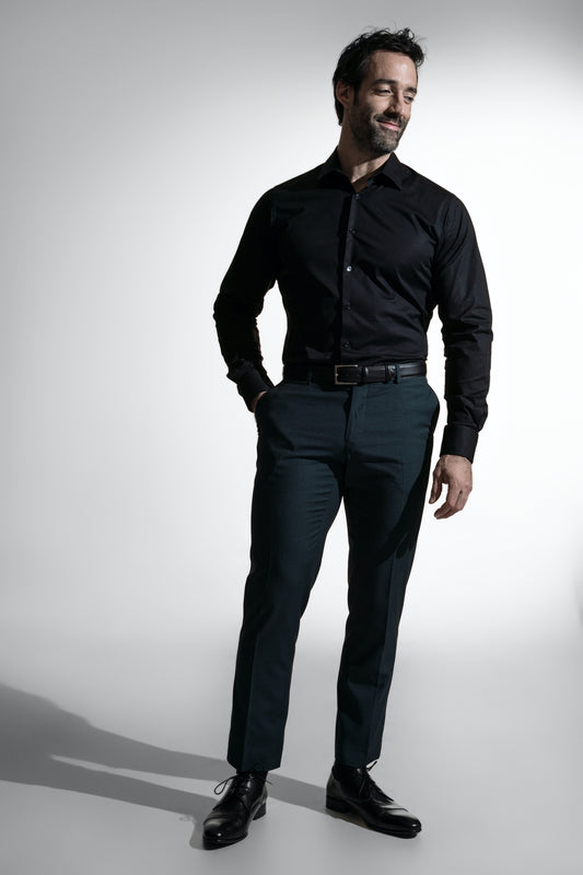 Stilfuld mand med skæg i en sort skjorte og mørke bukser, udstråler selvtillid mod en enkel baggrund