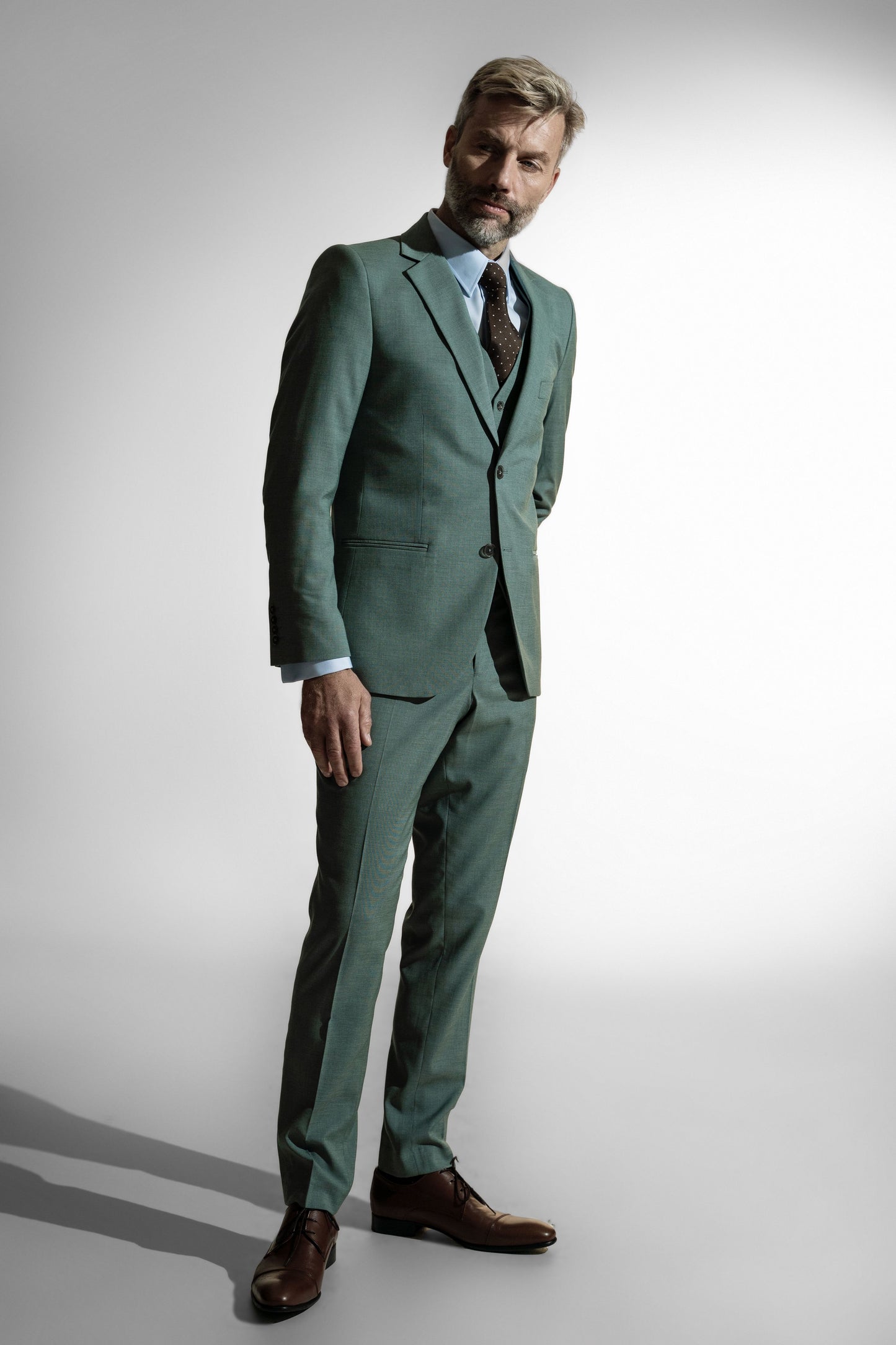 Stilfuld moden mand i en moderne pastelgrøn skræddersyet jakkesæk, brun prikket slips og lædersko, sat mod en bleg baggrund