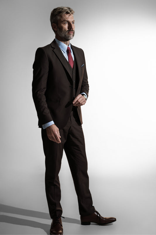 Moden mand med sølvfarvet hår i en klassisk brun skræddersyet jakkesæk, rød slips, og lædersko, poserer mod en lys baggrund