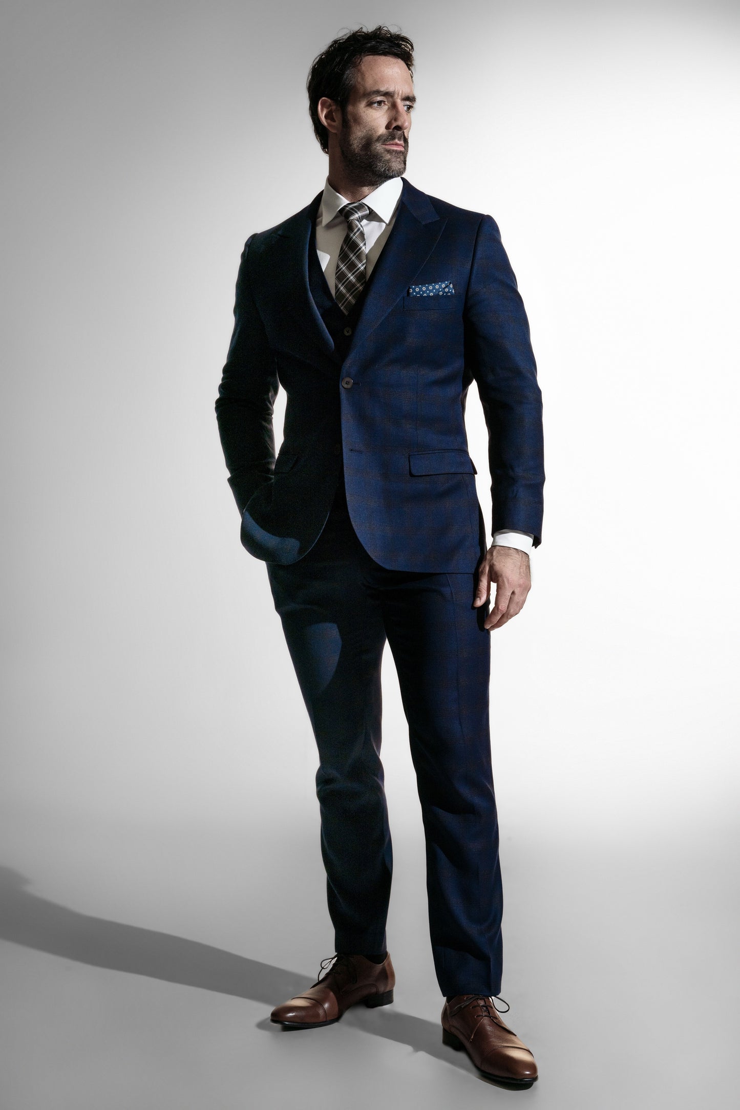 Stilfuld mand i en dybblå skræddersyet jakkesæk med sort slips og lommetørklæde, ser eftertænksom ud mod en hvid baggrund