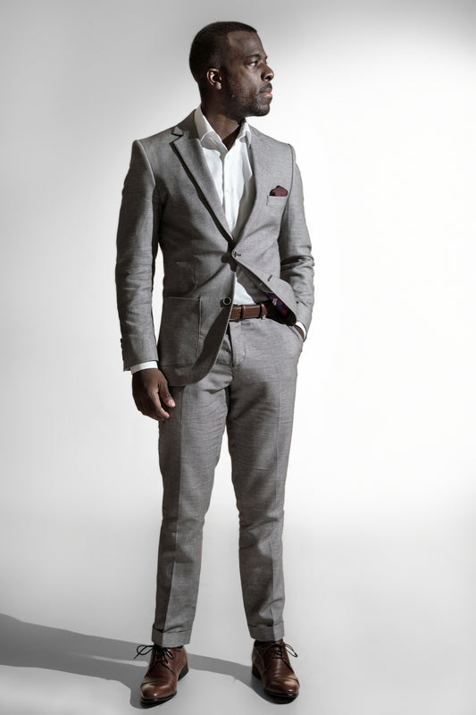 Tankefuld afroamerikansk mand i en lys grå skræddersyet jakkesæk, hvid skjorte og brun bælte, stående mod en hvid baggrund